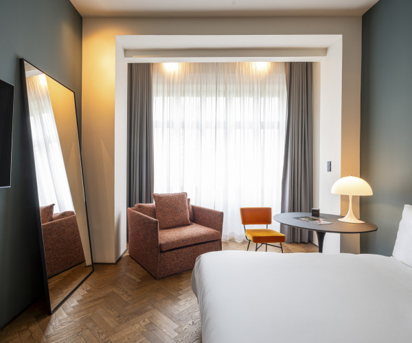 Modern hotel room with a bed, armchair, orange chair, round table, lamp, large mirror, TV, and floor-to-ceiling window with curtains.
