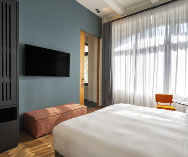 Modern hotel room with a large bed, wall-mounted TV, orange chair, and large window with sheer white curtains.