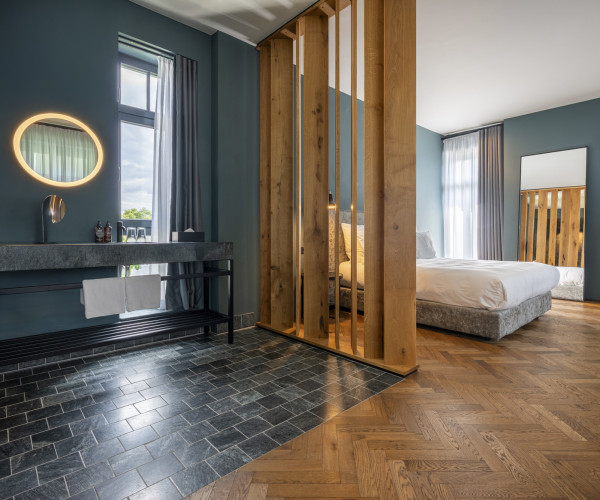 Modern hotel room with a partitioned bathroom area, large mirrors, wood flooring, and a bed with natural light from tall windows.