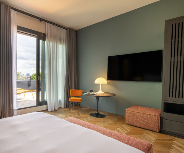 Modern hotel room with a large bed, wall-mounted TV, small table, orange chair, and balcony with outdoor seating visible through glass doors.