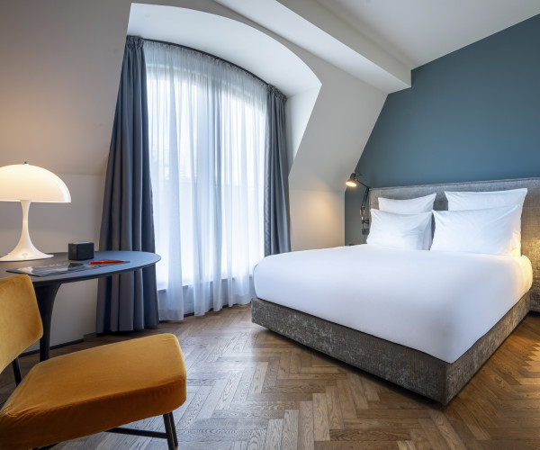 Modern hotel room with a double bed, blue accent wall, desk, lamp, two yellow chairs, and large window with curtains.