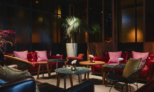 Eine Lounge mit roten und dunklen Ledersofas, runden Holz- und Marmortischen, gemusterten Kissen, Topfpflanzen und gedämpfter Beleuchtung.