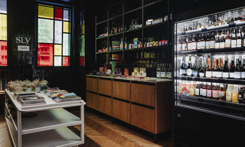 Ein kleiner Laden mit Regalen mit verschiedenen Lebensmitteln, einem gläsernen Kühlschrank mit Getränken und einem Wagen mit Büchern und Gläsern in der Nähe von Buntglasfenstern.