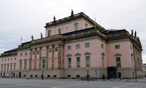 Ein großes, blassrosa neoklassizistisches Gebäude mit Statuen auf dem Dach, das auf einem leeren, gepflasterten Platz unter einem bewölkten Himmel steht.