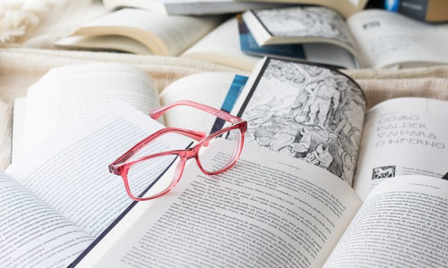 Eine rosa Brille liegt auf einem offenen Buch, umgeben von anderen offenen Büchern, einige mit Text und einige mit Illustrationen.