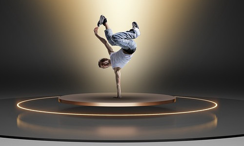 Ein Breakdancer balanciert auf einer Hand auf einer runden Bühne, umgeben von leuchtend gelben Ringen und dramatischer Beleuchtung.