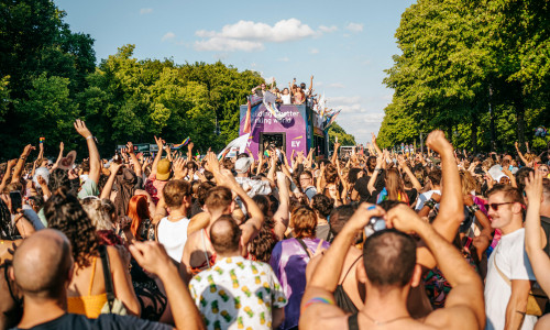 Eine große Menschenmenge feiert bei einer Pride-Veranstaltung im Freien und streckt unter einem sonnigen Himmel ihre Hände in Richtung eines farbenfrohen Festwagens.